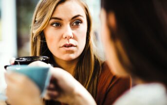 A woman with long brown hair is holding a cup and appears to be engaged in a conversation. The focus is on her expression, as she listens intently. Another person in the foreground is blurred, also holding a cup.