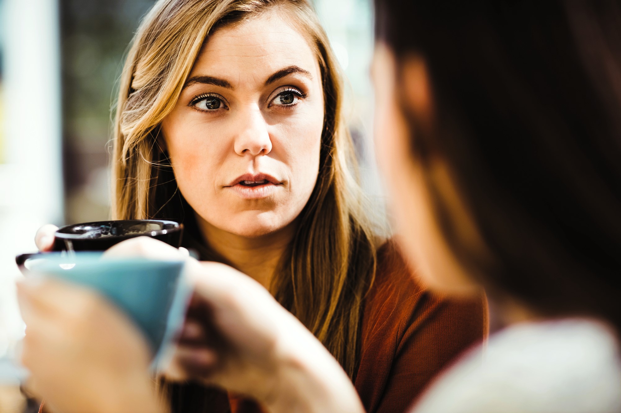 A woman with long brown hair is holding a cup and appears to be engaged in a conversation. The focus is on her expression, as she listens intently. Another person in the foreground is blurred, also holding a cup.