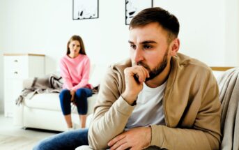 A man in a beige jacket sits on a couch, appearing contemplative, with his hand on his chin. In the background, a woman in a pink sweater sits on another part of the couch, looking concerned. The room is bright with simple decor.