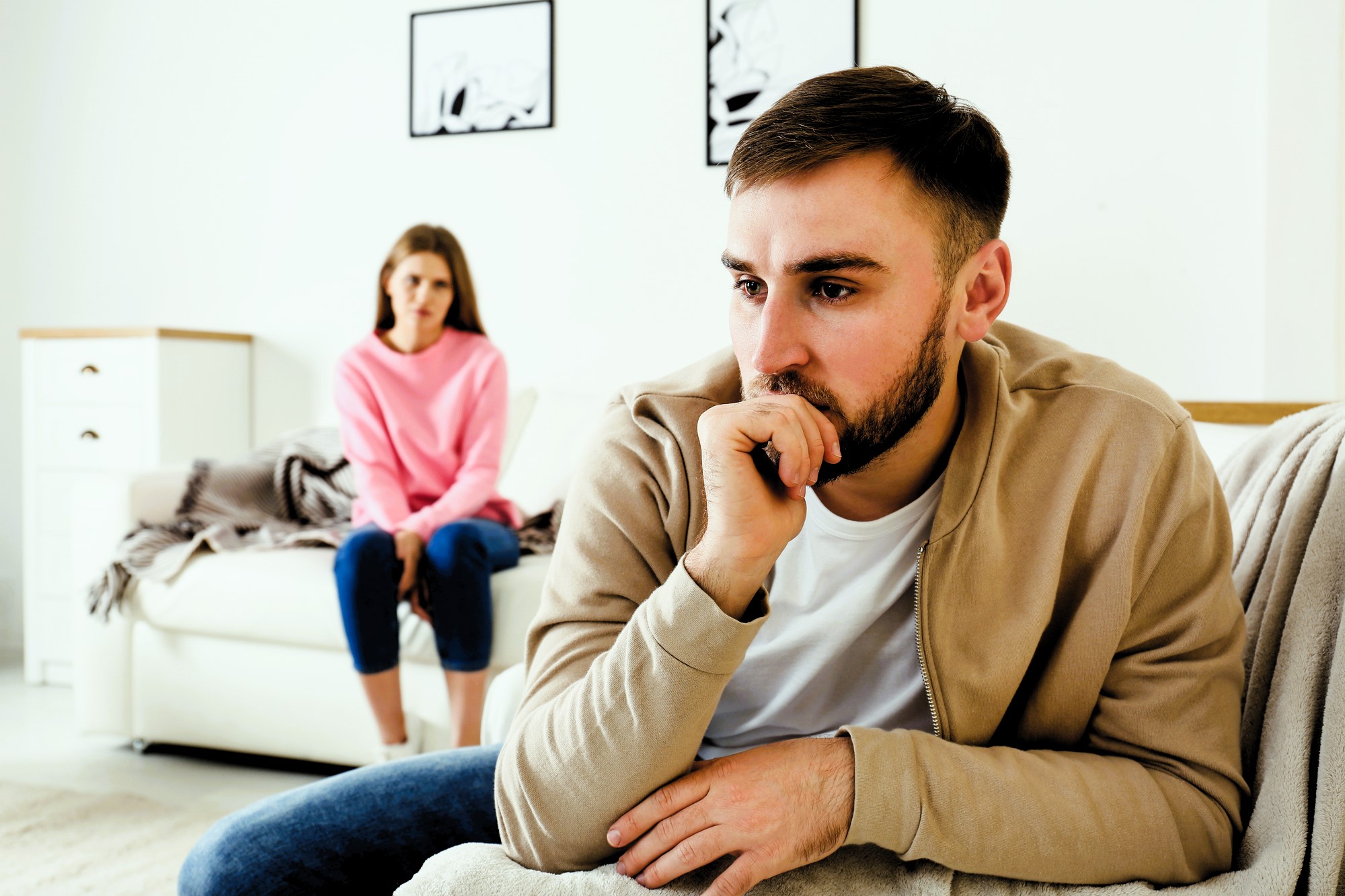 A man in a beige jacket sits on a couch, appearing contemplative, with his hand on his chin. In the background, a woman in a pink sweater sits on another part of the couch, looking concerned. The room is bright with simple decor.