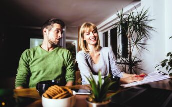 A man and woman sit at a table with paperwork. The man in a green sweater looks at the woman, who is smiling and wearing a gray top. A laptop, coffee mugs, cookies, and potted plants are on the table. The room is softly lit with plants in the background.