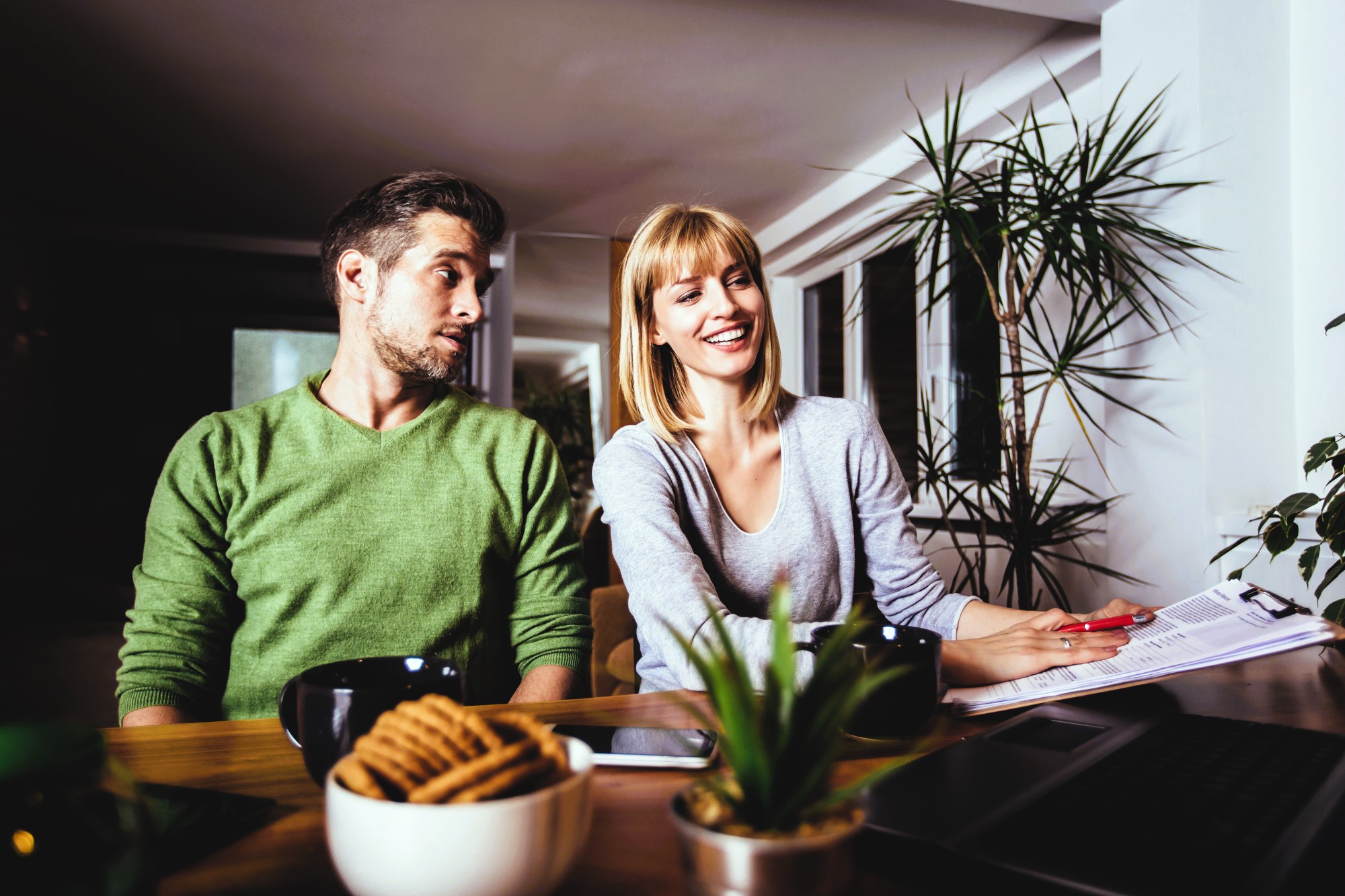 A man and woman sit at a table with paperwork. The man in a green sweater looks at the woman, who is smiling and wearing a gray top. A laptop, coffee mugs, cookies, and potted plants are on the table. The room is softly lit with plants in the background.