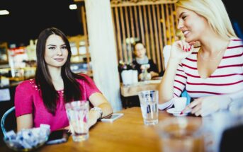 Two women sit at a wooden table in a cafe, with drinks and smartphones in front of them. One is wearing a pink top and the other a white top. They are smiling and engaged in conversation. The background shows a blurred cafe interior with people.