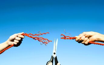 Two hands are pulling apart bright red rope against a clear blue sky, while a pair of scissors in the center appears to be cutting the strands, causing them to fray on either side.