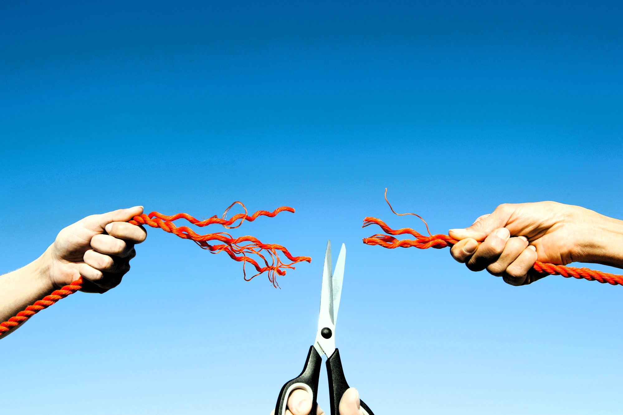 Two hands are pulling apart bright red rope against a clear blue sky, while a pair of scissors in the center appears to be cutting the strands, causing them to fray on either side.