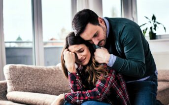 A woman sitting on a couch appears upset, with her head in her hand. A man beside her, wearing a green sweater, leans in to comfort her with his arm around her shoulders. They are indoors, near large windows with blurred outdoor view.