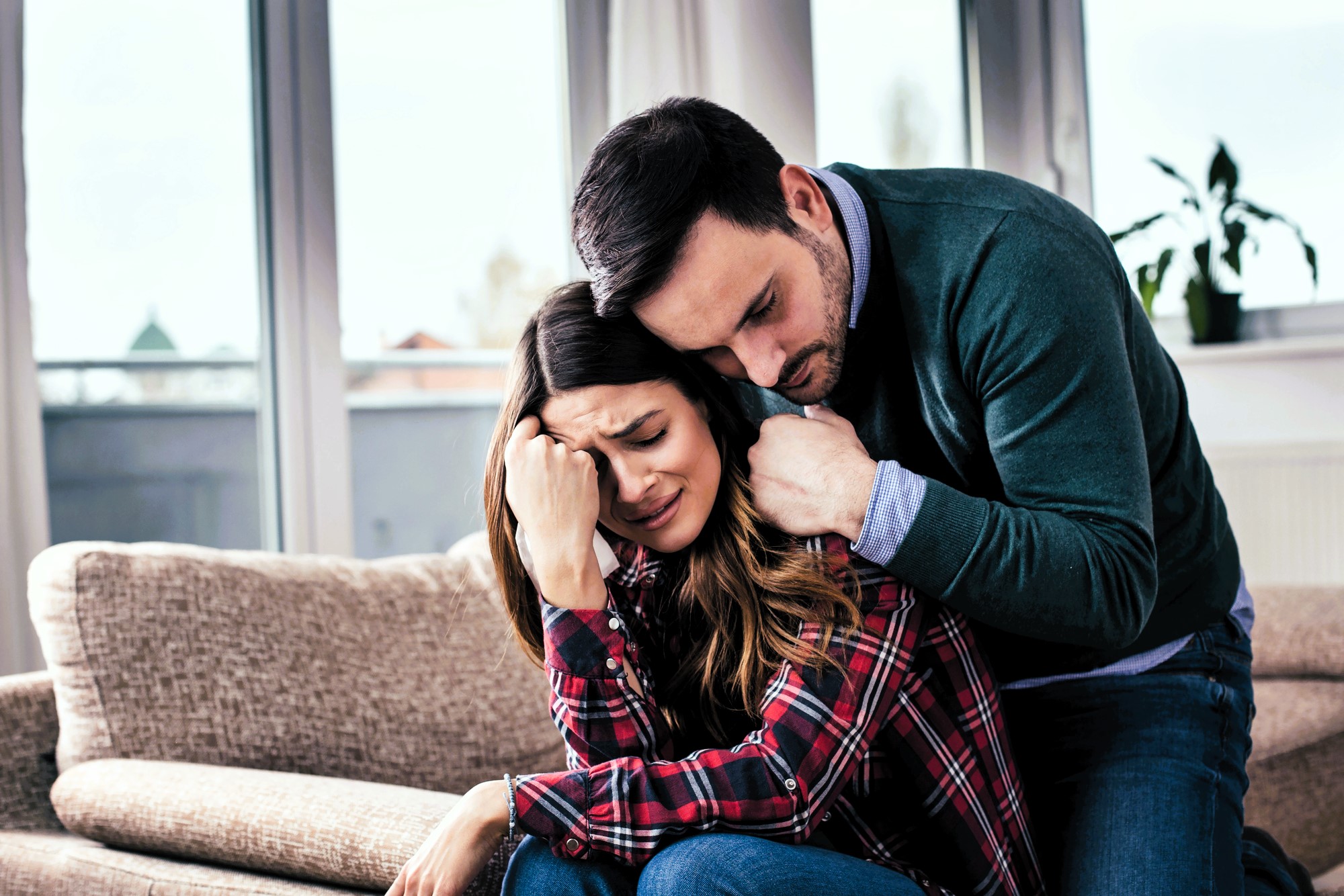 A woman sitting on a couch appears upset, with her head in her hand. A man beside her, wearing a green sweater, leans in to comfort her with his arm around her shoulders. They are indoors, near large windows with blurred outdoor view.