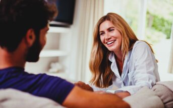 A woman with long hair, wearing a light shirt, smiles and leans forward on a couch while speaking to a man with a beard and dark shirt. They are in a cozy, sunlit living room with curtains and greenery outside.