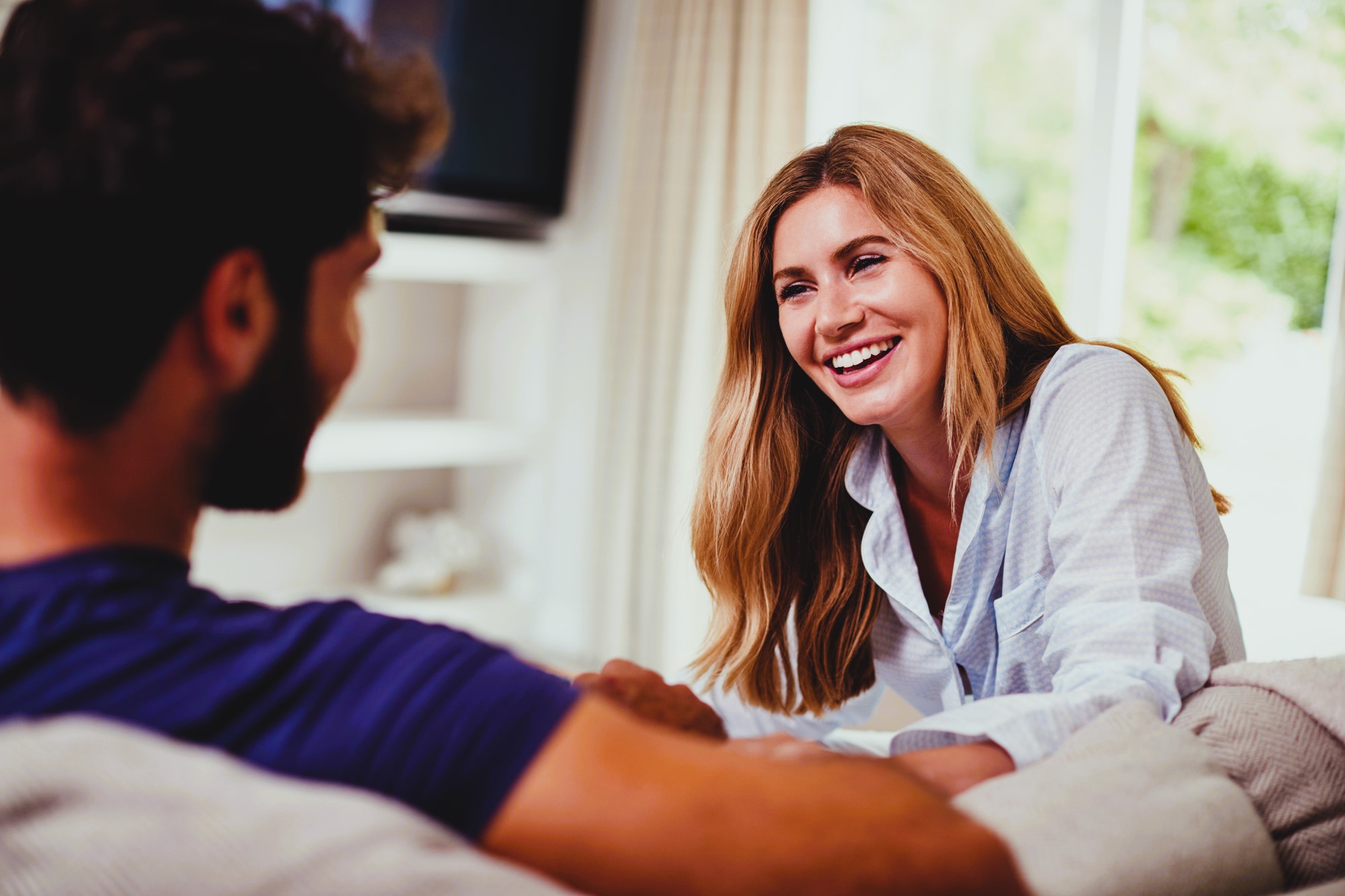 A woman with long hair, wearing a light shirt, smiles and leans forward on a couch while speaking to a man with a beard and dark shirt. They are in a cozy, sunlit living room with curtains and greenery outside.