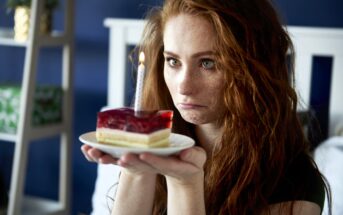 A woman with long red hair looks at a small cake slice with a lit candle on top, which she is holding on a white plate. She appears to be pouting. The background is a blurred interior setting with a bed.