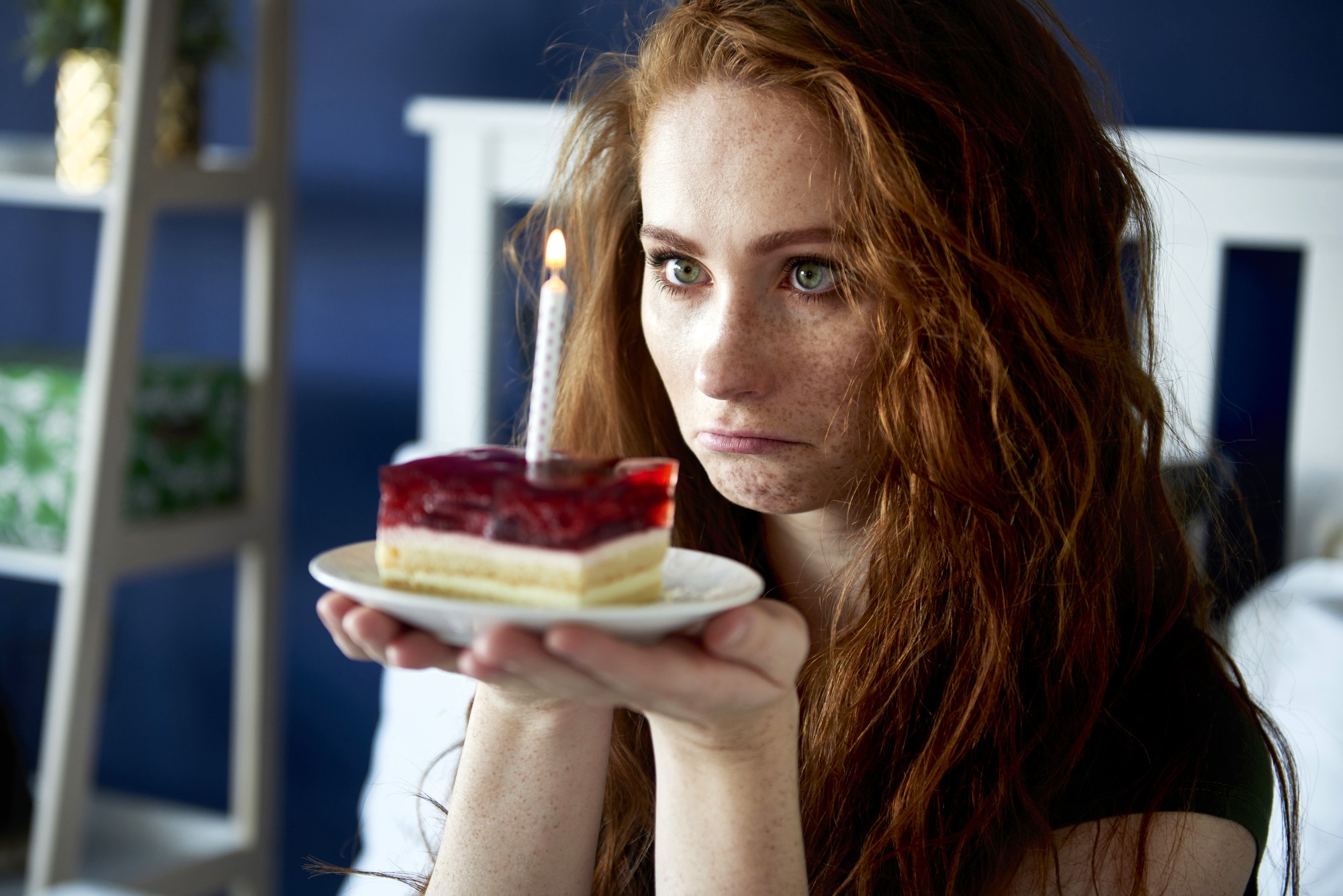 A woman with long red hair looks at a small cake slice with a lit candle on top, which she is holding on a white plate. She appears to be pouting. The background is a blurred interior setting with a bed.