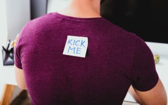 A person wearing a purple shirt has a sticky note on their back with the words "KICK ME" written in blue. The person is sitting at a desk, facing away from the camera.