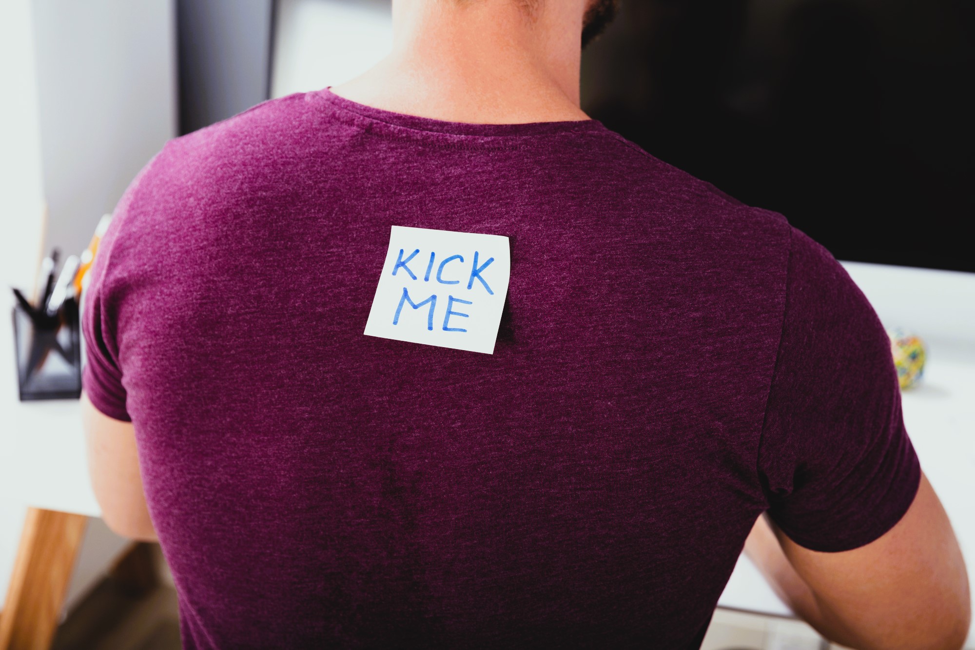 A person wearing a purple shirt has a sticky note on their back with the words "KICK ME" written in blue. The person is sitting at a desk, facing away from the camera.