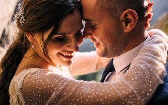 A couple embraces closely, smiling with foreheads touching, in a sunlit setting. The woman wears a white, embellished dress and has decorative hairpins, while the man is in a suit. Their expressions are joyful and serene.