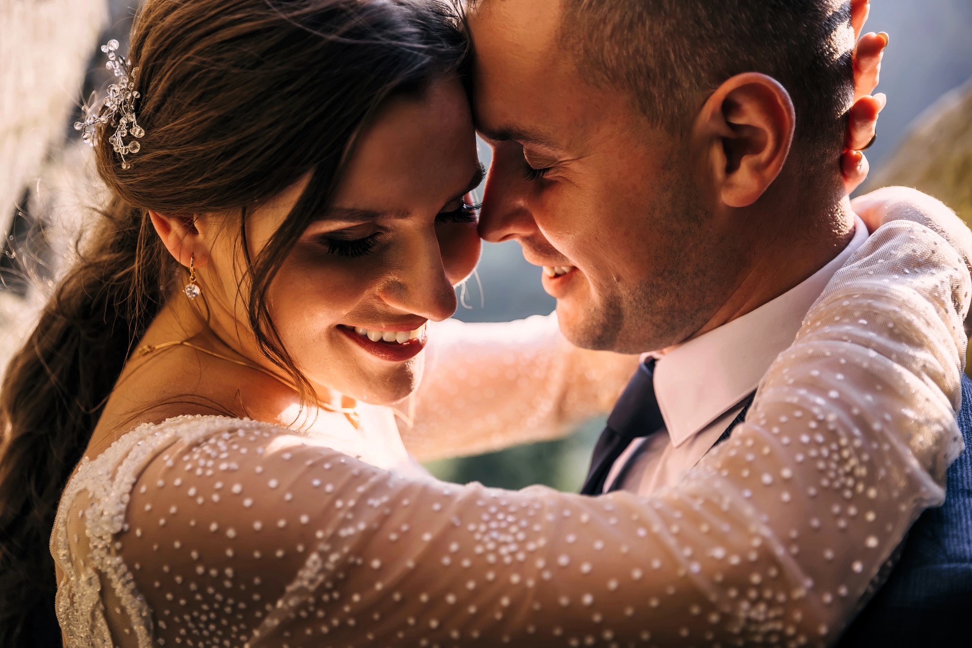 A couple embraces closely, smiling with foreheads touching, in a sunlit setting. The woman wears a white, embellished dress and has decorative hairpins, while the man is in a suit. Their expressions are joyful and serene.