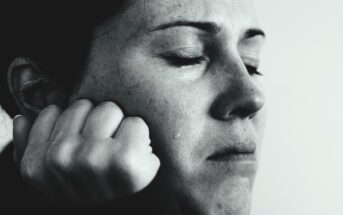 Close-up of a person with eyes closed, appearing distressed or thoughtful. A single tear runs down the cheek, and their hand is clenched near the chin. The image is in black and white, conveying an emotional or contemplative mood.