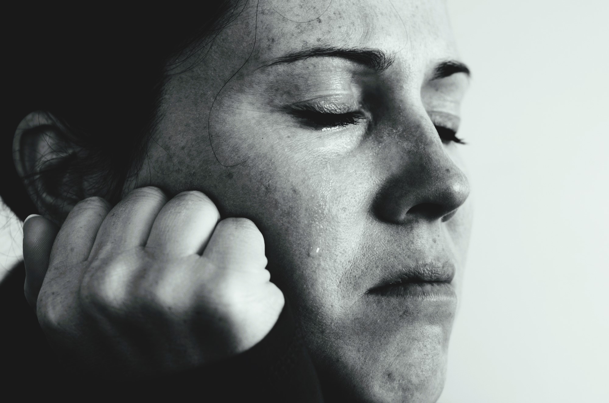 Close-up of a person with eyes closed, appearing distressed or thoughtful. A single tear runs down the cheek, and their hand is clenched near the chin. The image is in black and white, conveying an emotional or contemplative mood.