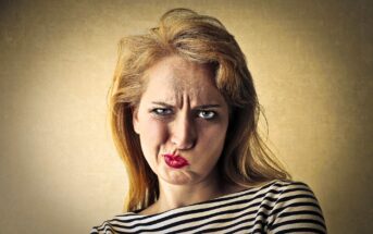 Woman with long blond hair wearing a striped shirt makes a displeased face with furrowed brow and pursed lips against a textured tan background.