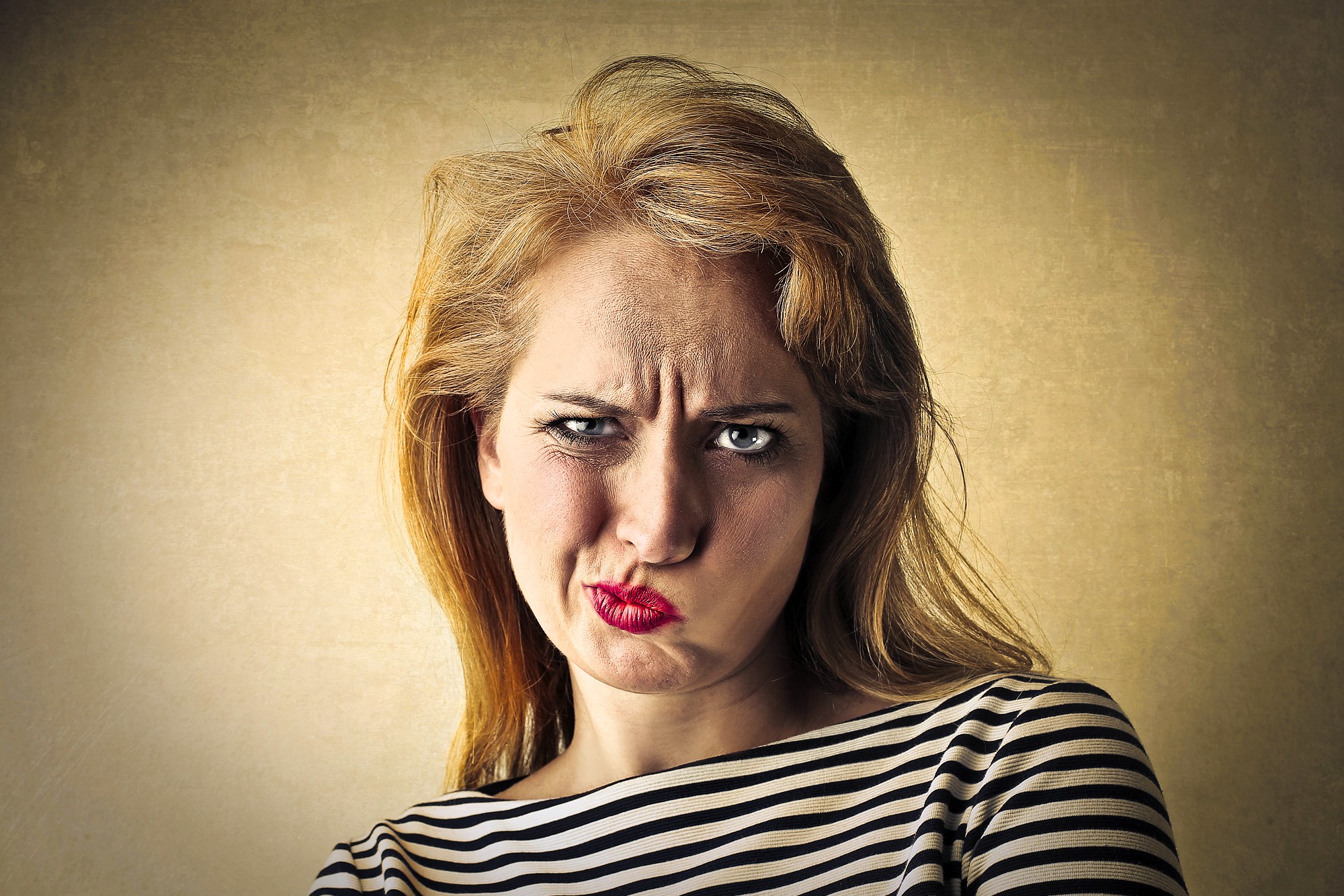 Woman with long blond hair wearing a striped shirt makes a displeased face with furrowed brow and pursed lips against a textured tan background.