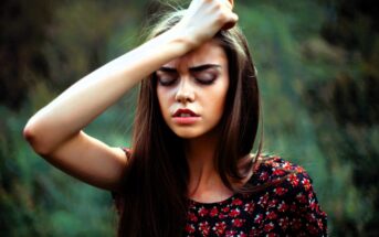 A woman with long brown hair stands outdoors with her eyes closed, wearing a floral-patterned dress. Her right arm is raised, resting her forehead on her hand. The background is blurred greenery.