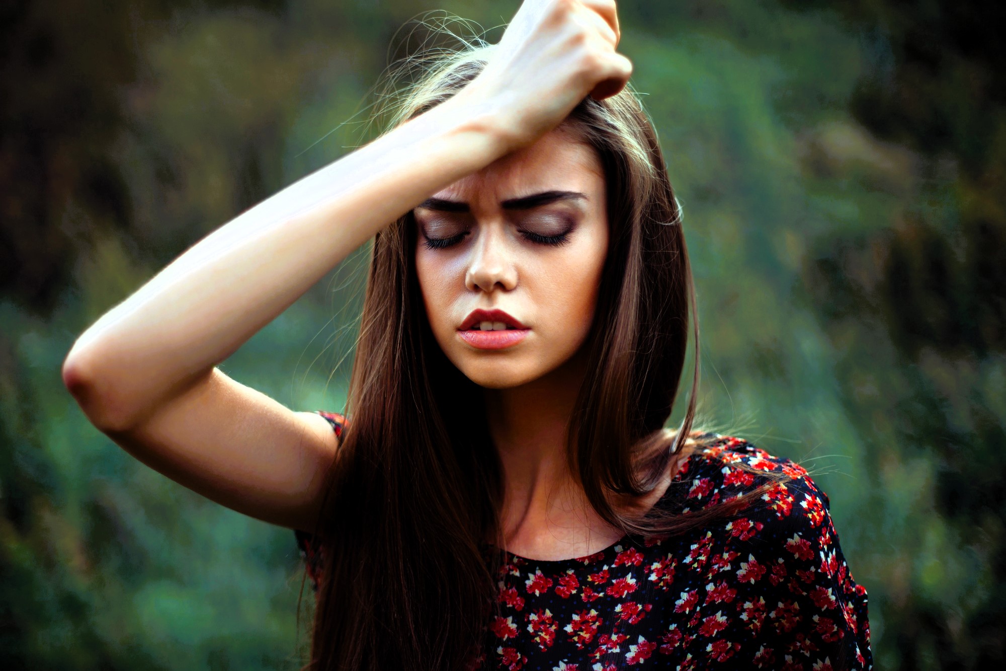 A woman with long brown hair stands outdoors with her eyes closed, wearing a floral-patterned dress. Her right arm is raised, resting her forehead on her hand. The background is blurred greenery.