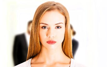 A woman with long blonde hair and red lipstick looks directly at the camera against a white background. Two blurred figures in business attire stand behind her.