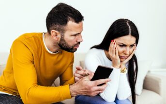 A man in a yellow sweater shows a smartphone to a woman in a white sweater. She looks worried and rests her chin on her hand. They are sitting on a beige sofa against a plain white background.