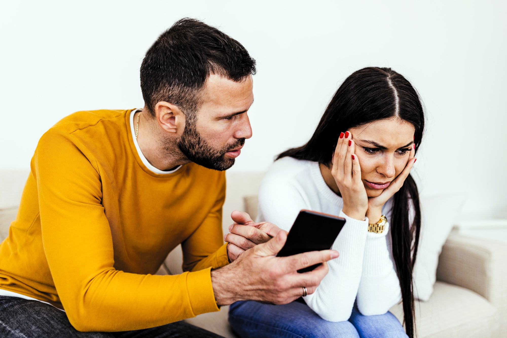 A man in a yellow sweater shows a smartphone to a woman in a white sweater. She looks worried and rests her chin on her hand. They are sitting on a beige sofa against a plain white background.