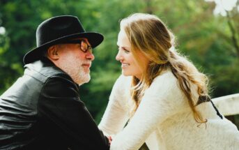 A man in a black hat and leather jacket leans towards a smiling woman with long blond hair in a light sweater. They are outdoors with green foliage in the background.