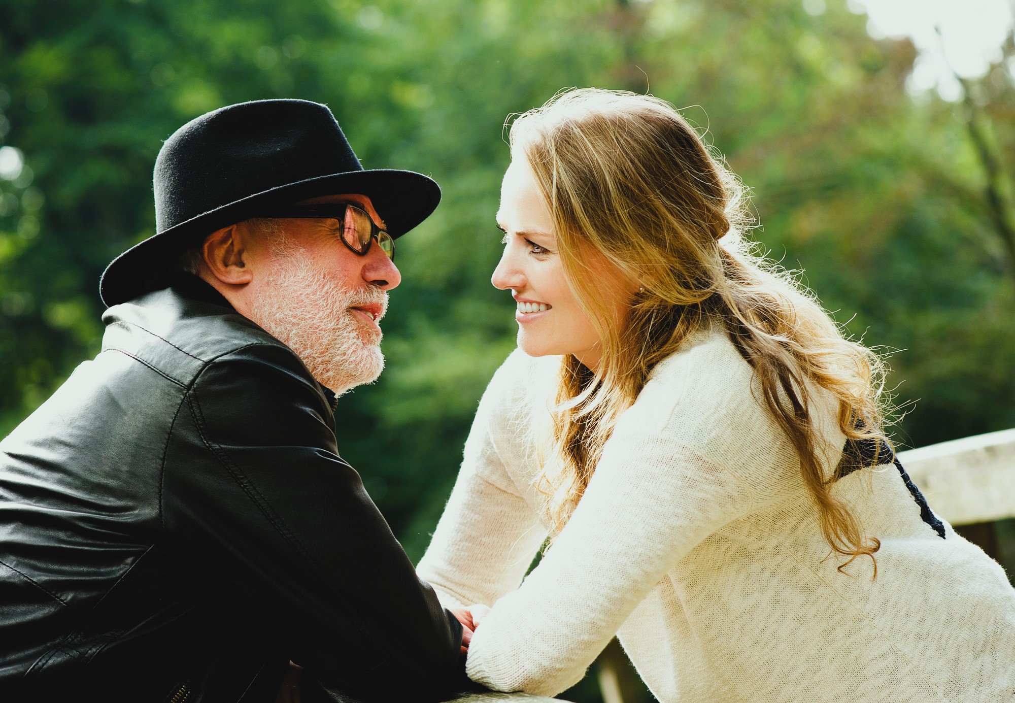 A man in a black hat and leather jacket leans towards a smiling woman with long blond hair in a light sweater. They are outdoors with green foliage in the background.