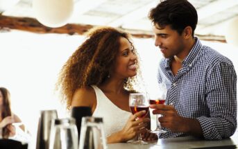 A couple stands at a bar, smiling and making eye contact while holding drinks. The woman has curly hair and wears a white dress, and the man is in a checkered shirt. They appear to be enjoying a relaxed conversation.