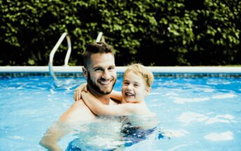 A man and a child are smiling and embracing while standing in a swimming pool. The water is clear, and a lush green hedge is visible in the background.