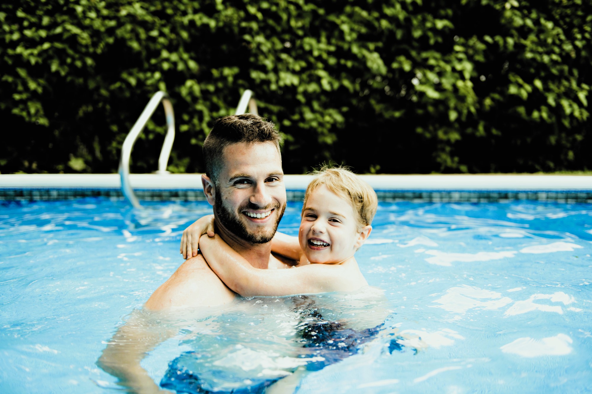 A man and a child are smiling and embracing while standing in a swimming pool. The water is clear, and a lush green hedge is visible in the background.