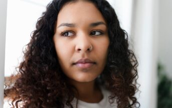 A person with long, curly hair and a nose ring looks pensively to the side. They are indoors with soft lighting and a blurred background.