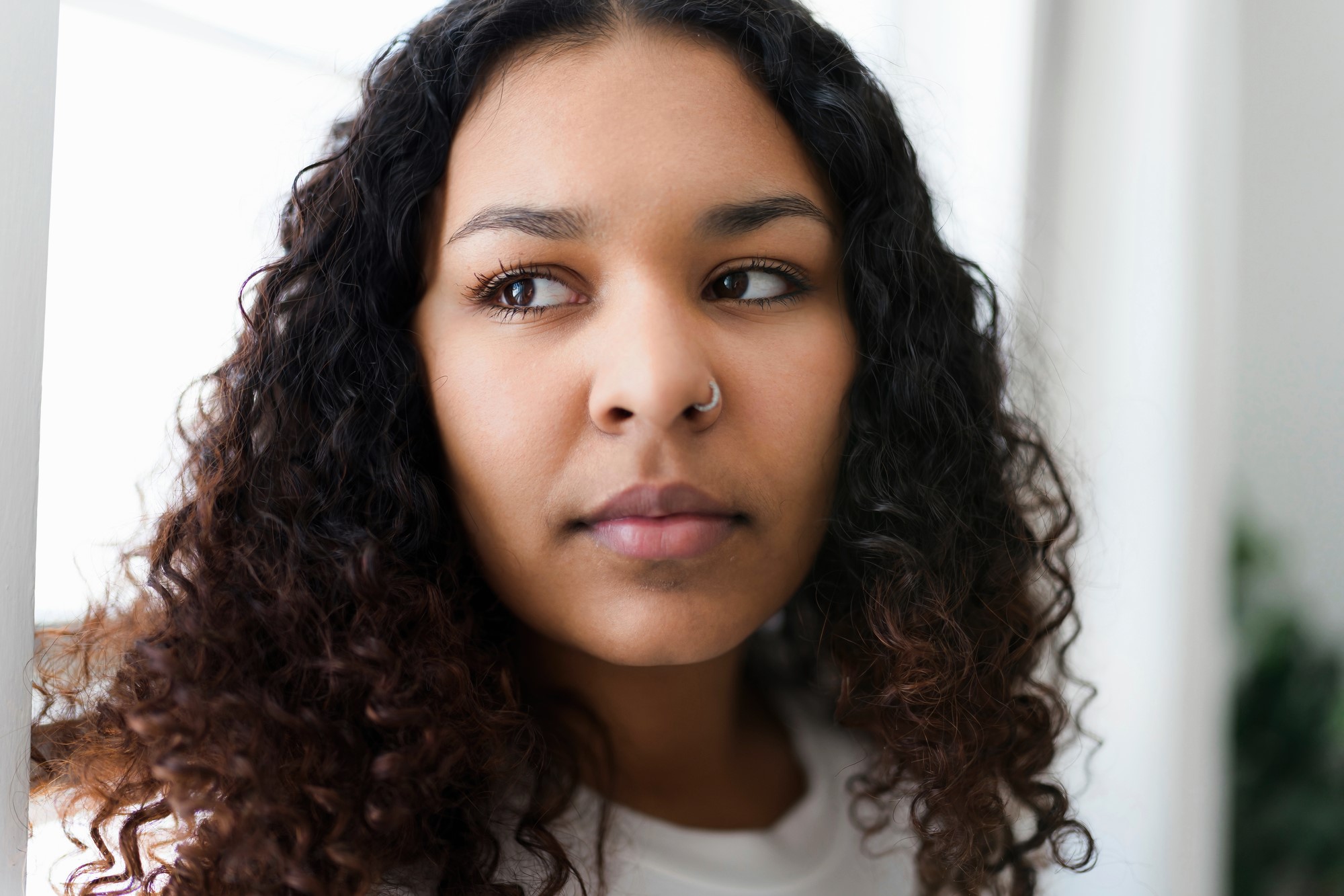 A person with long, curly hair and a nose ring looks pensively to the side. They are indoors with soft lighting and a blurred background.