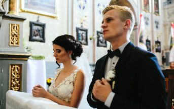 A bride and groom stand side by side in a church, both looking contemplative. The bride, in a lacy white dress, has her hands clasped, while the groom, in a dark suit and bow tie, holds his hands to his chest. Ornate decorations are visible in the background.