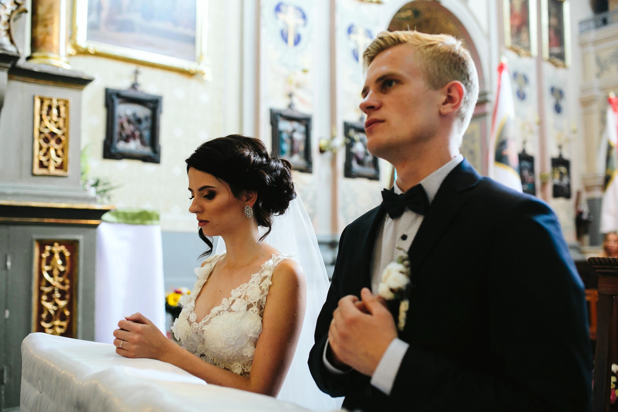 A bride and groom stand side by side in a church, both looking contemplative. The bride, in a lacy white dress, has her hands clasped, while the groom, in a dark suit and bow tie, holds his hands to his chest. Ornate decorations are visible in the background.