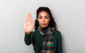 A woman with long, dark curly hair stands against a gray background. She is wearing a green sweater and a multicolored scarf. Her right hand is raised with her palm facing outward, conveying a gesture to stop or halt.