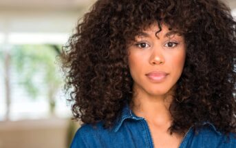 A woman with curly hair looks at the camera with a neutral expression. She is wearing a blue denim top. The background is softly blurred, suggesting an indoor setting.