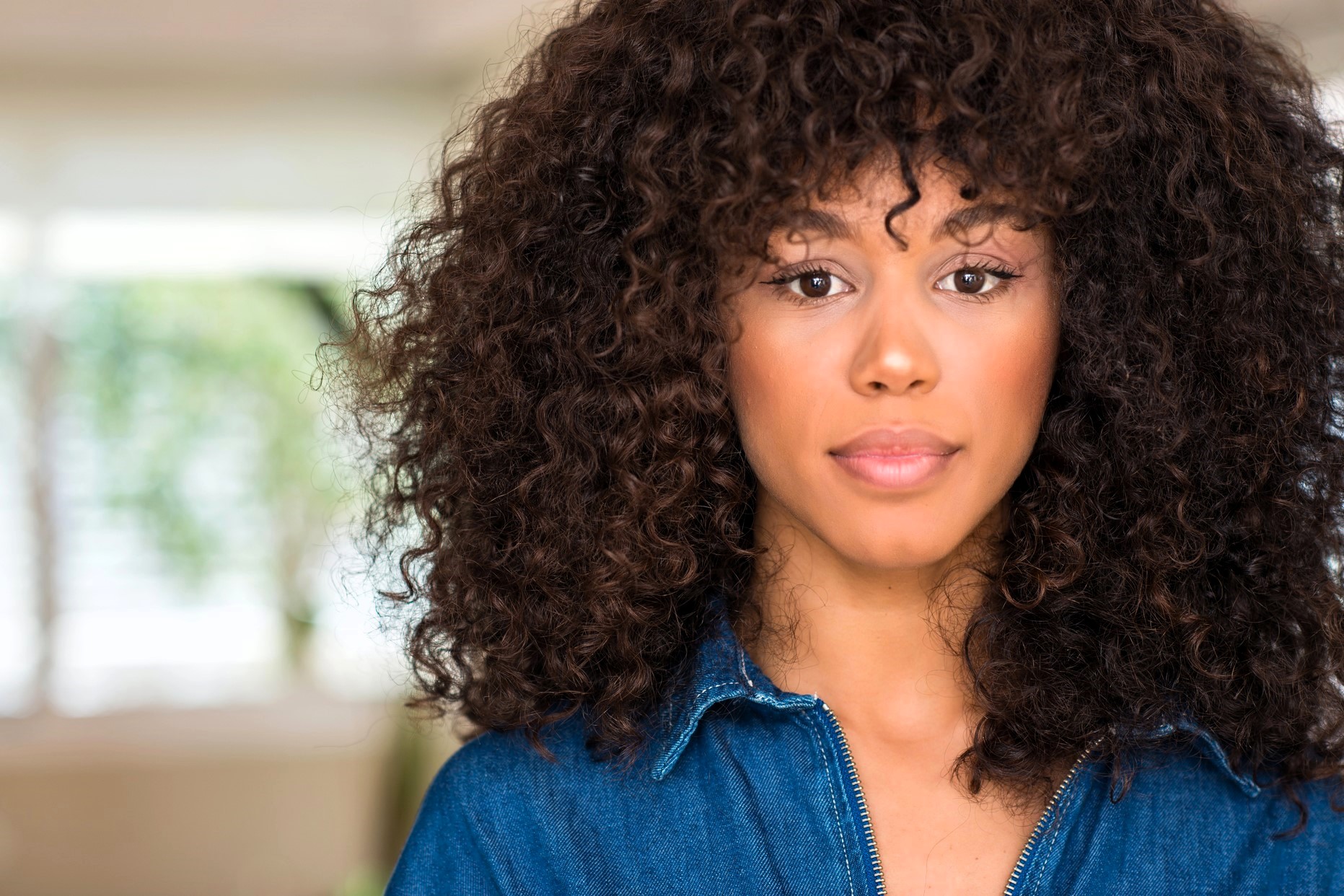 A woman with curly hair looks at the camera with a neutral expression. She is wearing a blue denim top. The background is softly blurred, suggesting an indoor setting.