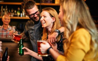 A group of friends laughing and enjoying drinks at a bar. A man with glasses has his arm around a smiling woman holding a beer mug. Another woman in a yellow outfit holds a bottle. The background shows shelves with various bottles.