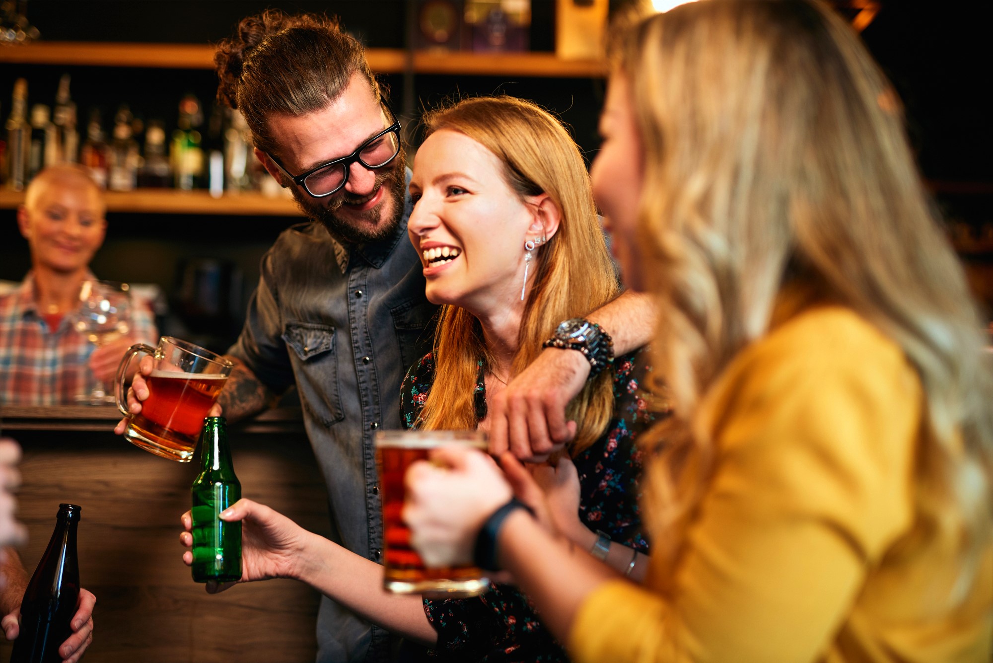 A group of friends laughing and enjoying drinks at a bar. A man with glasses has his arm around a smiling woman holding a beer mug. Another woman in a yellow outfit holds a bottle. The background shows shelves with various bottles.