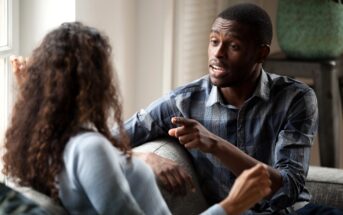 A man and woman are sitting on a couch, engaged in a conversation. The man, wearing a plaid shirt, gestures while speaking. The woman, with curly hair, is facing him, listening attentively. The setting is a well-lit room with a window in the background.