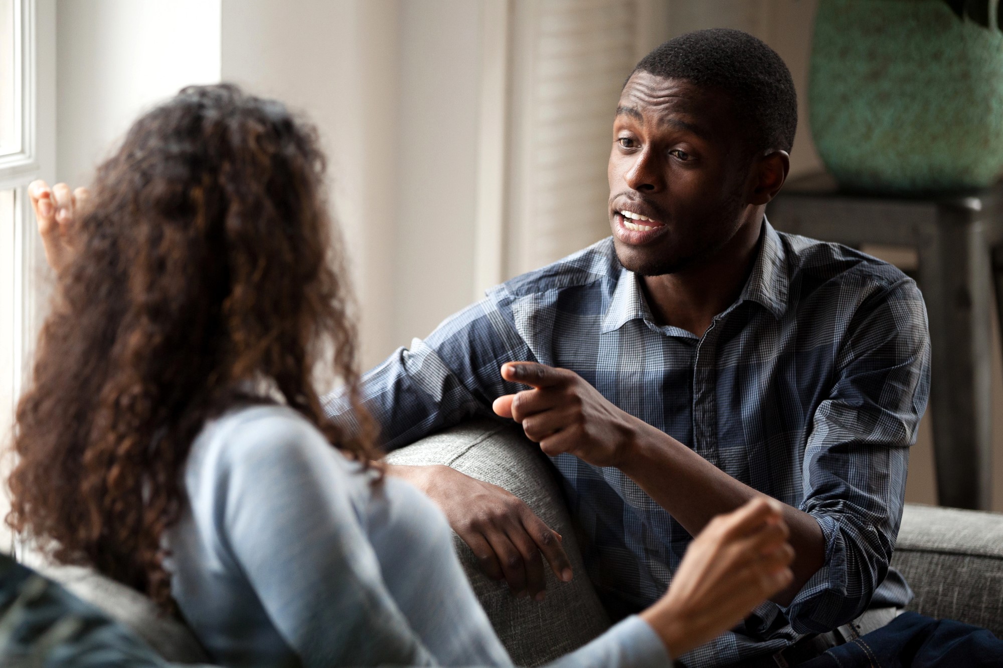 A man and woman are sitting on a couch, engaged in a conversation. The man, wearing a plaid shirt, gestures while speaking. The woman, with curly hair, is facing him, listening attentively. The setting is a well-lit room with a window in the background.