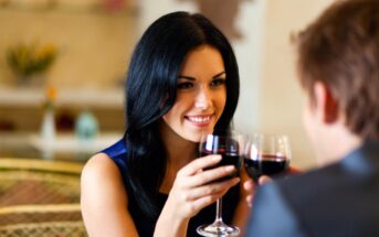 A woman with long black hair and a man, both holding glasses of red wine, sit across from each other in a cozy setting. The woman is smiling warmly as they toast. The background is softly blurred, creating an intimate atmosphere.