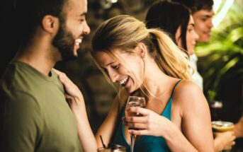 A group of people socializing at a party. A woman in a blue dress is laughing, holding a drink while talking to a man in a green shirt. Two people are in the background, holding drinks and smiling. There are plants and dim lighting.