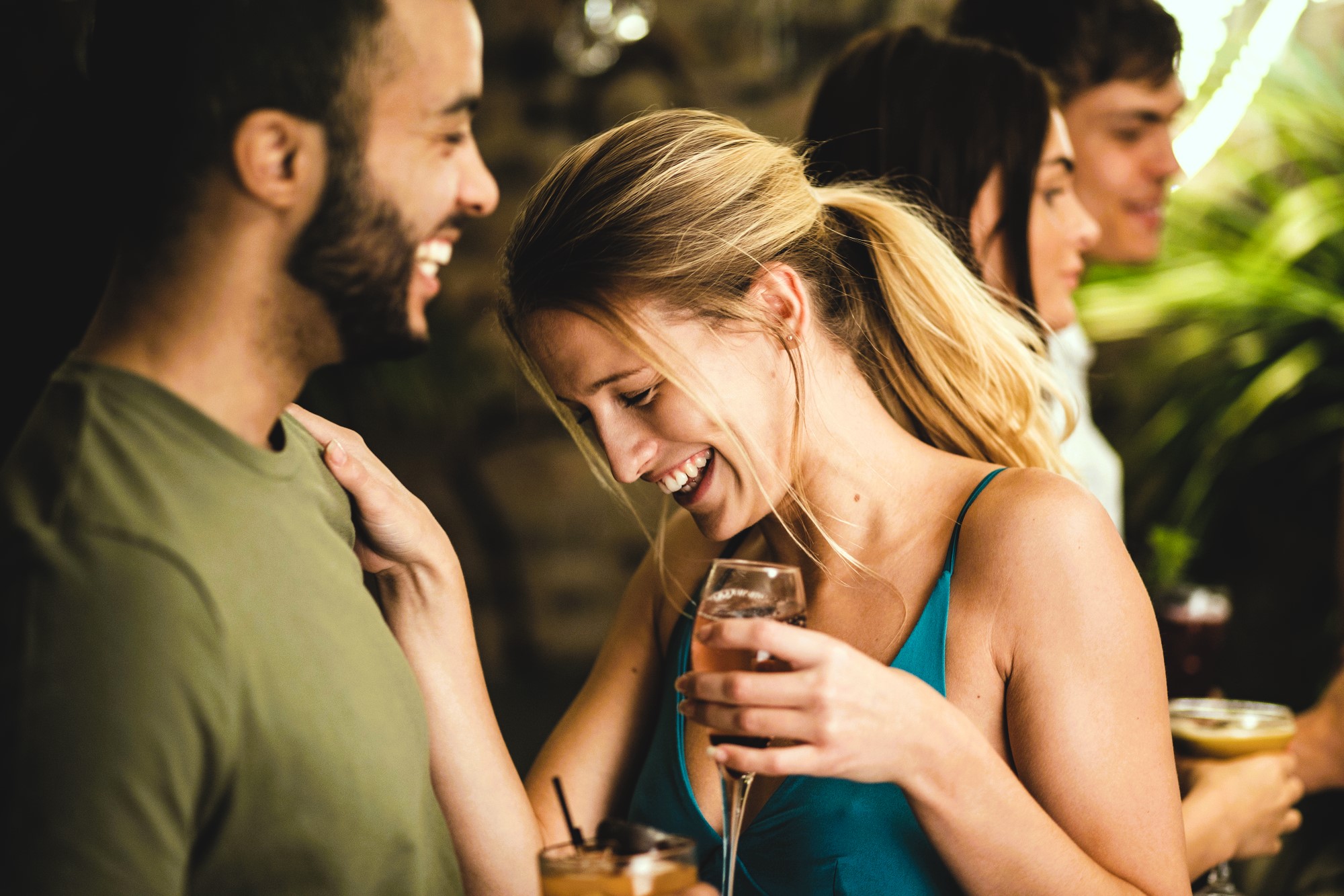 A group of people socializing at a party. A woman in a blue dress is laughing, holding a drink while talking to a man in a green shirt. Two people are in the background, holding drinks and smiling. There are plants and dim lighting.