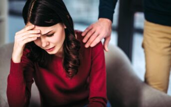 A woman in a red sweater sits with her head in her hand, appearing upset. A person's arm is gently placed on her shoulder, offering comfort. The background is softly blurred.