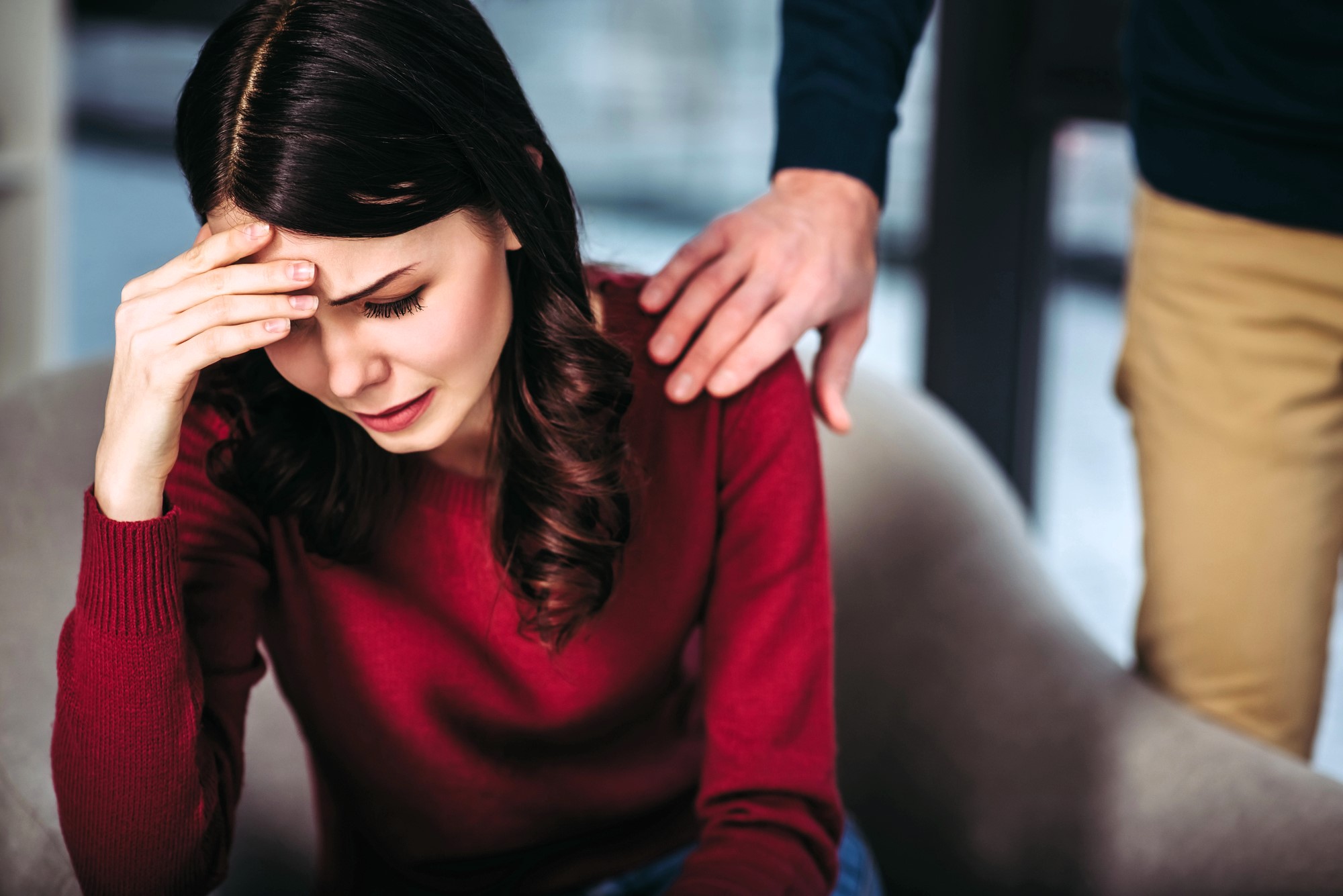 A woman in a red sweater sits with her head in her hand, appearing upset. A person's arm is gently placed on her shoulder, offering comfort. The background is softly blurred.