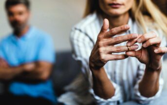 A woman sits in focus, looking down and removing a ring from her finger, symbolizing a separation or break-up. In the background, a man with crossed arms appears blurred, sitting on a couch. The scene conveys tension and emotional distance.